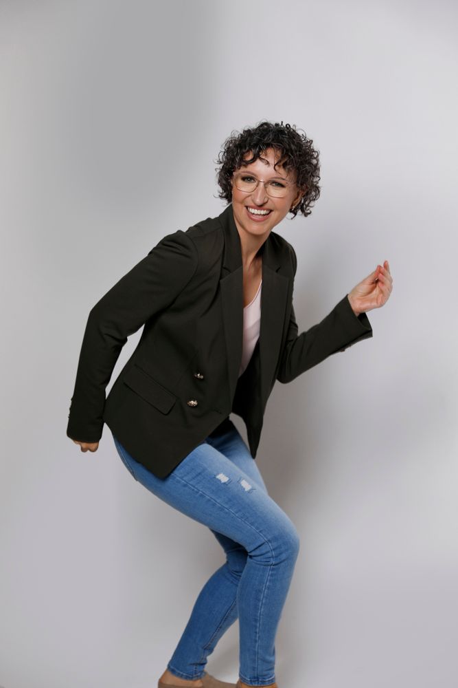 A curly-haired woman in a dark blazer and jeans dances playfully against a gray background.