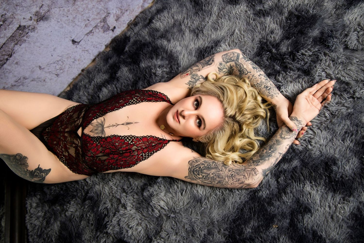 Tattooed blonde woman in red sequin bodysuit lying on gray fur rug.