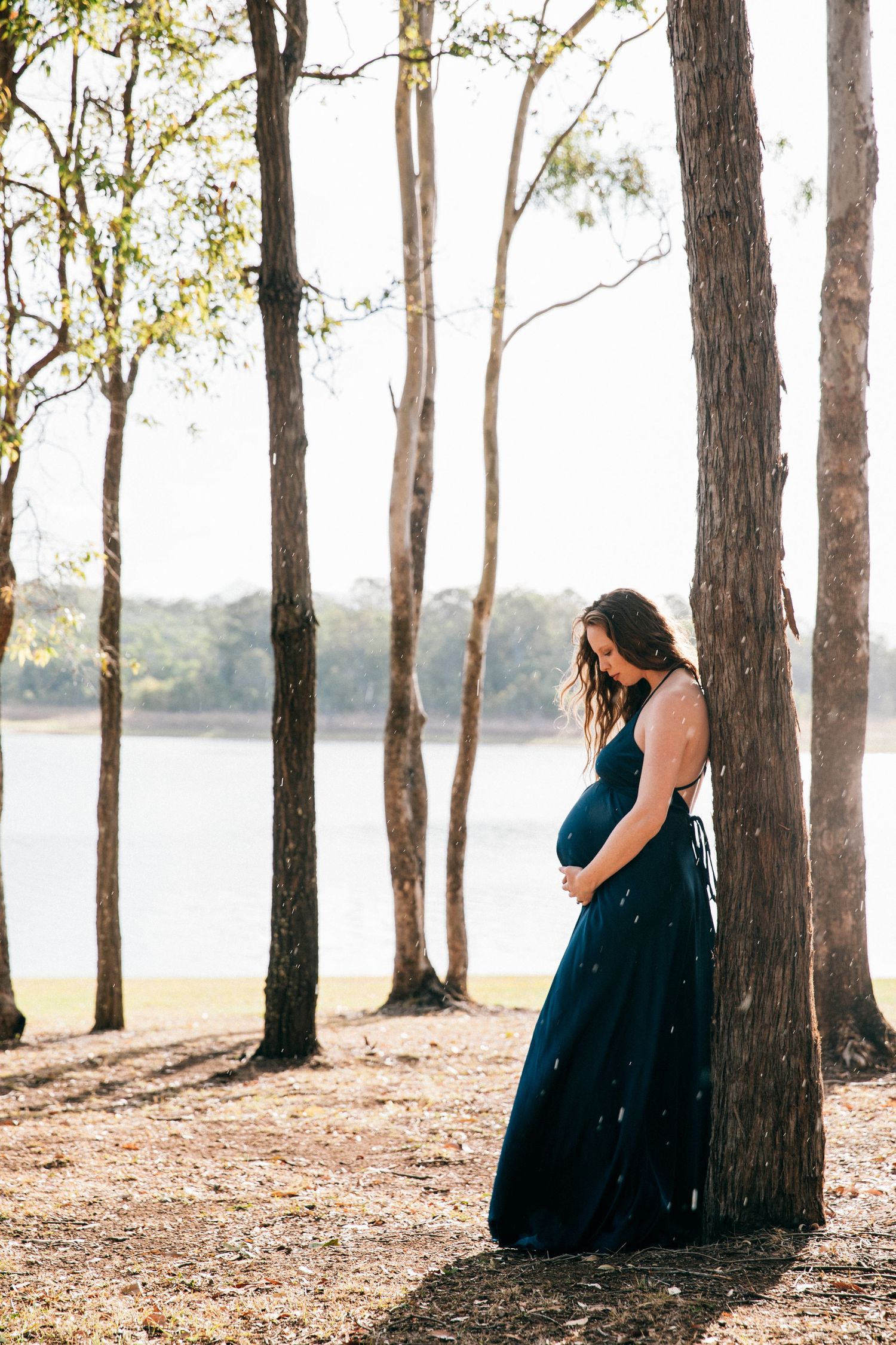 Pregnant woman in navy dress leaning against tree near lake during outdoor maternity photoshoot.
