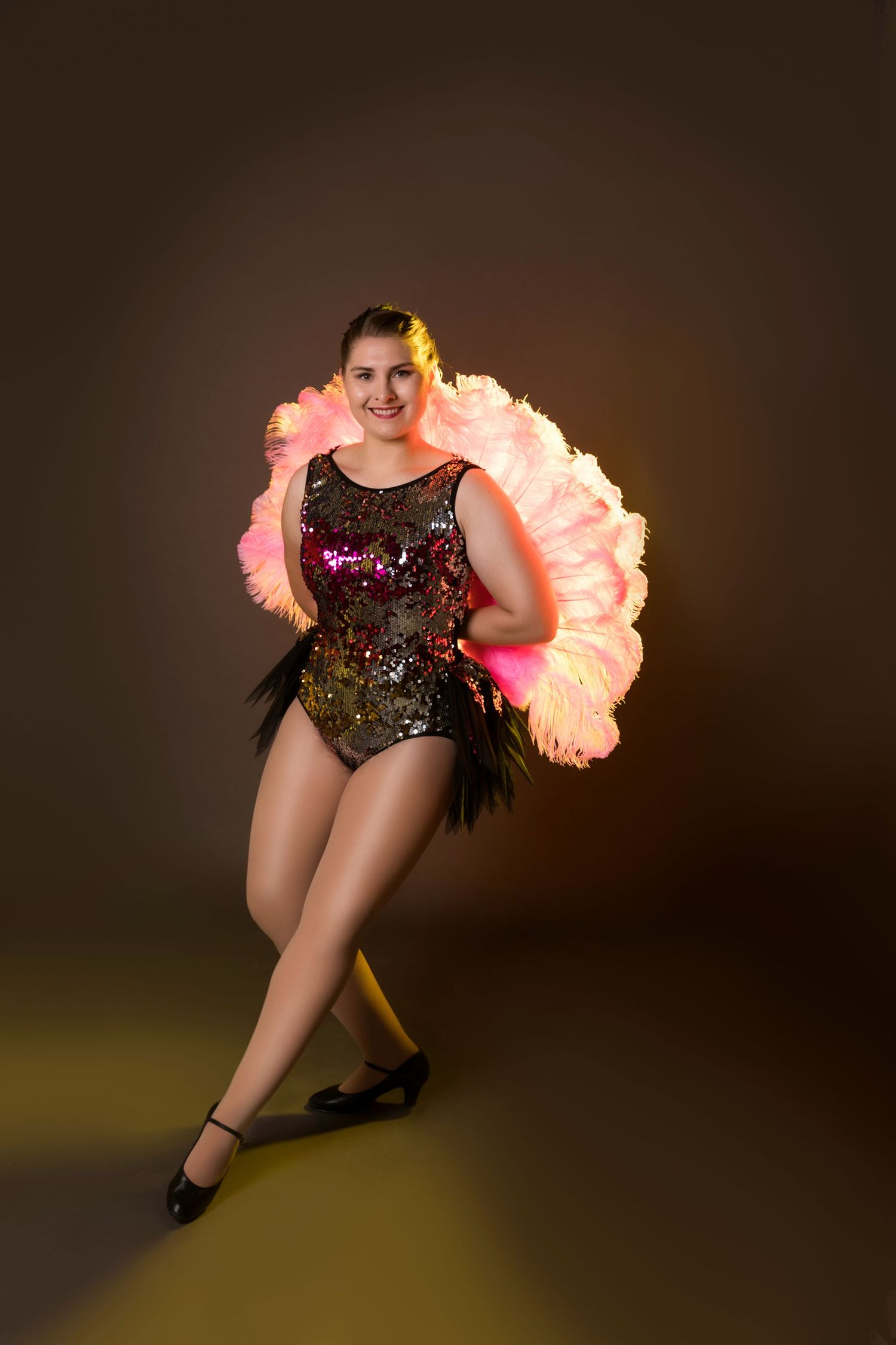 Smiling performer in sequined costume holds large pink feather fans on stage.