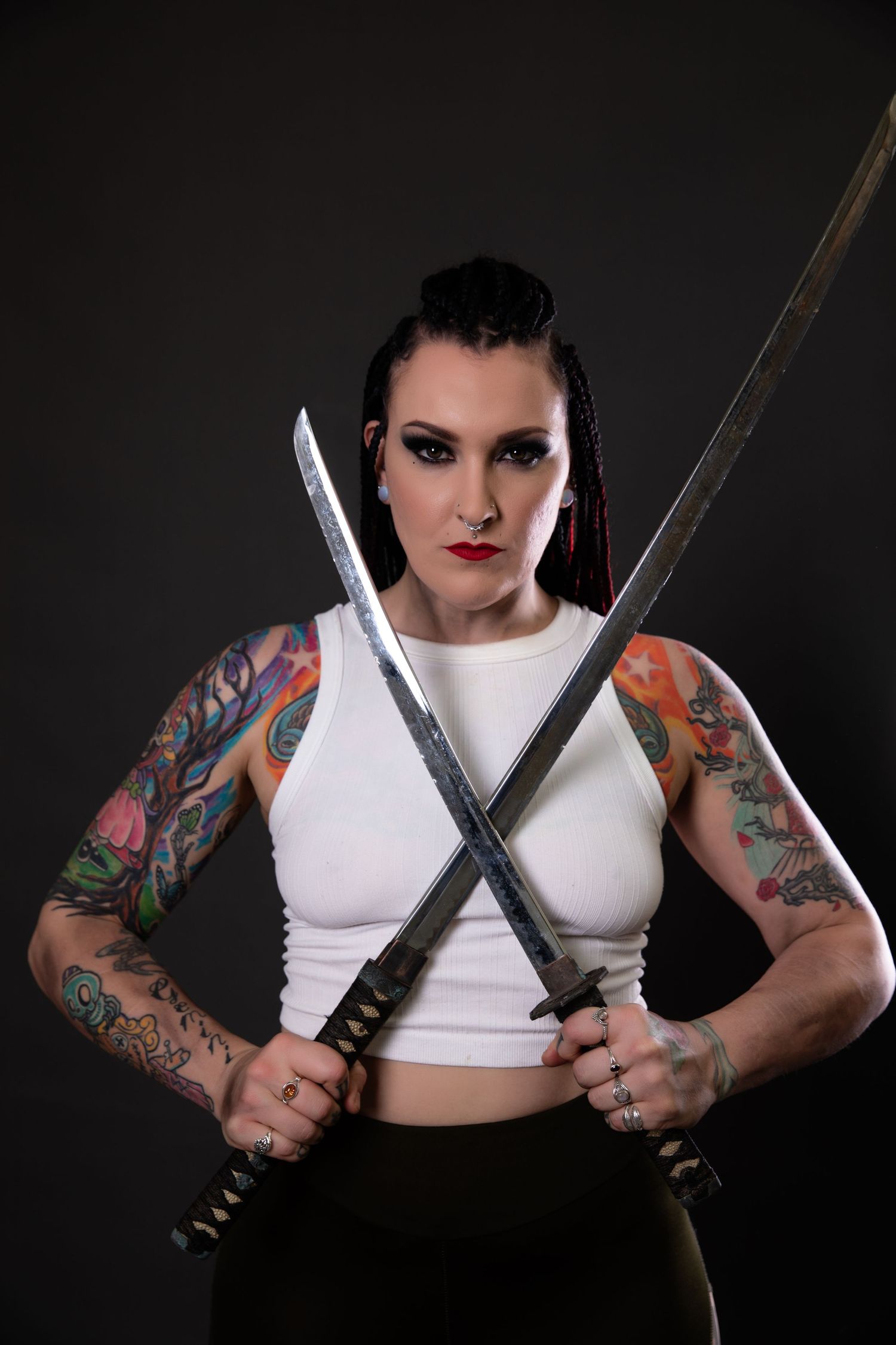 Tattooed woman in white crop top crosses two samurai swords in dramatic studio portrait.
