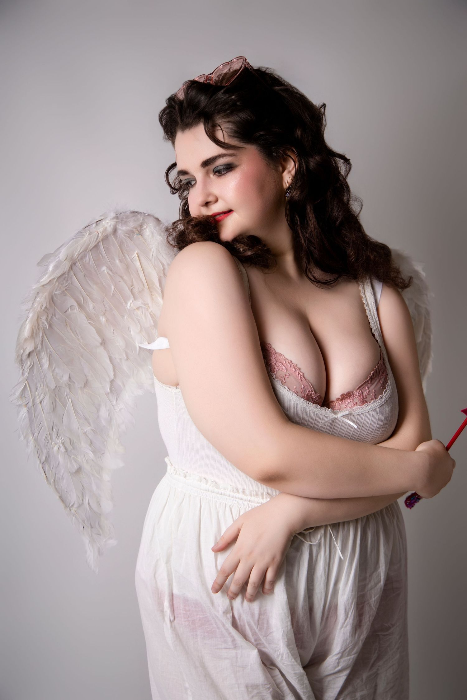 A plus-size woman dressed as an angel with white feather wings holding a red arrow in a studio.