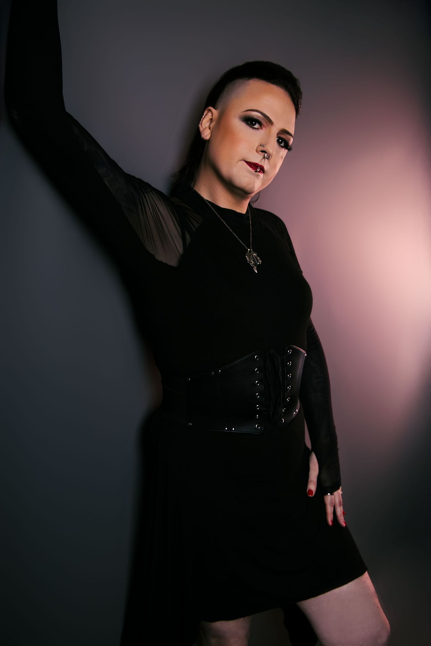 Gothic woman in black dress and corset belt posing against dark studio wall.
