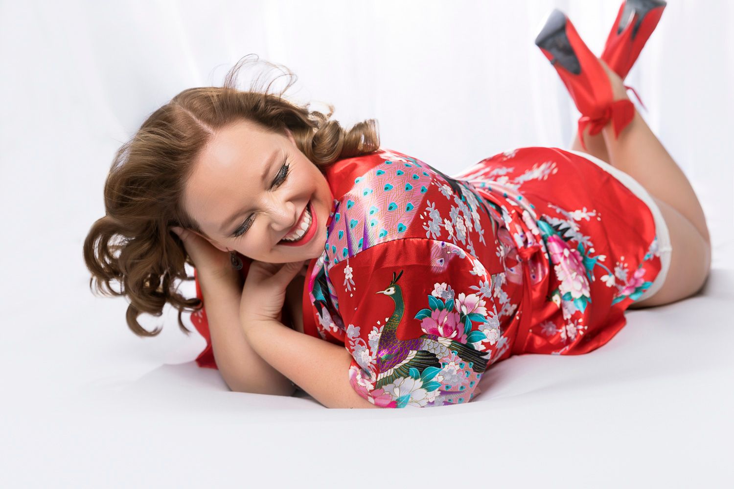 Woman in red floral kimono lying on white background smiling playfully at camera.
