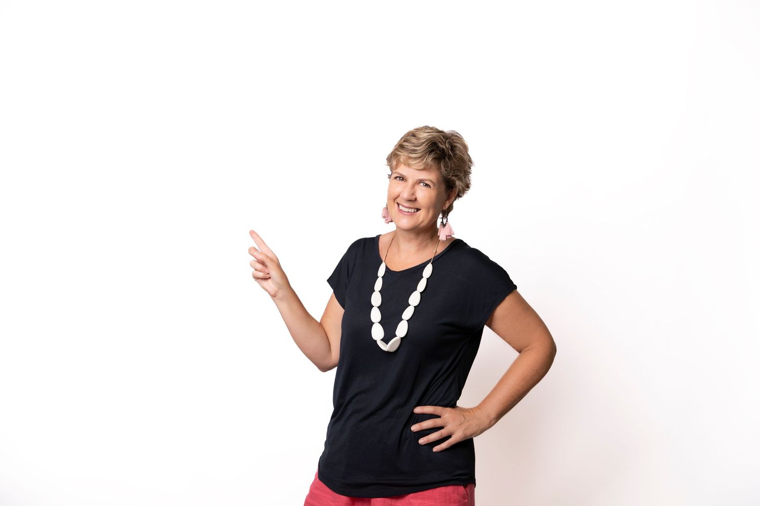 Smiling middle-aged woman in black top and white beaded necklace pointing left against white background.