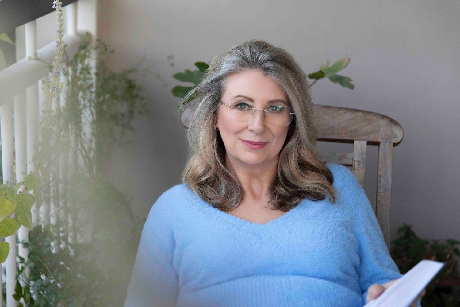 Elegant woman with gray hair in blue sweater sitting outdoors among green plants.