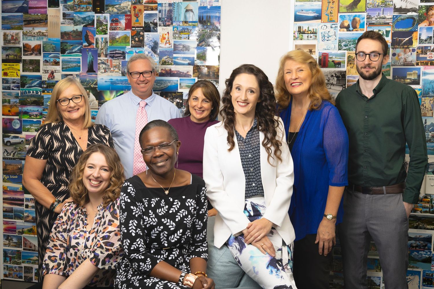 Group of nine smiling professionals posing together in front of a wall covered in colorful postcards.