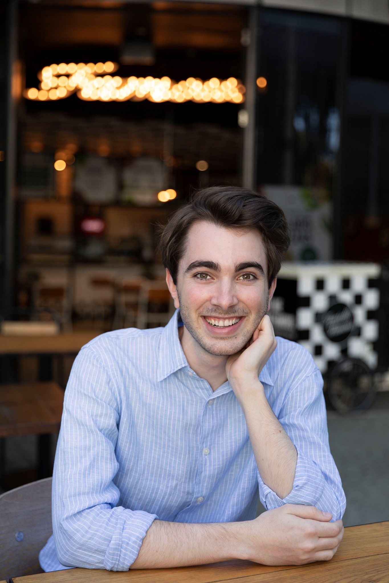 Smiling young man in blue shirt seated at outdoor cafe table with bokeh lights in background.