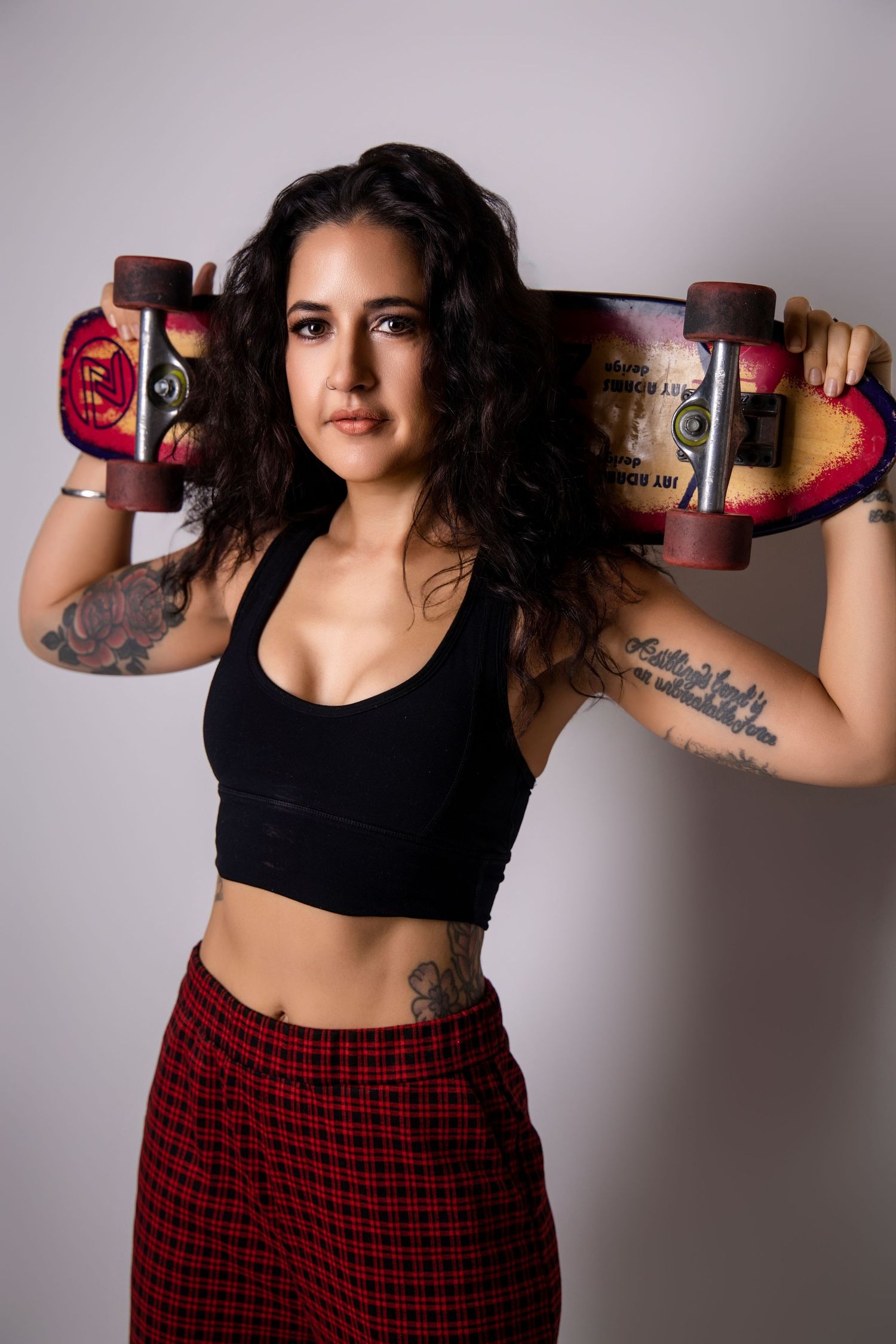 Tattooed brunette woman holding a skateboard behind her shoulders wearing a black crop top and plaid skirt.