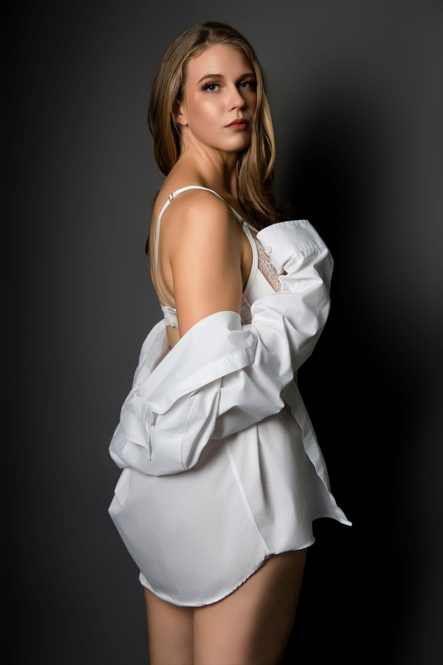 A blonde woman in a white oversized shirt and lingerie poses against a dark background.