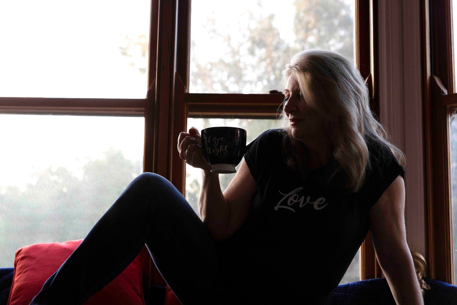 Woman relaxing by window holding coffee mug wearing black Love t-shirt in cozy setting.