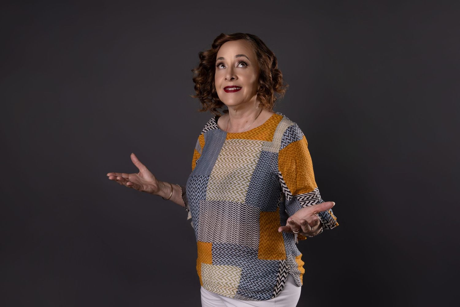 Expressive woman in colorful patchwork sweater gesturing with both hands against dark gray studio background.