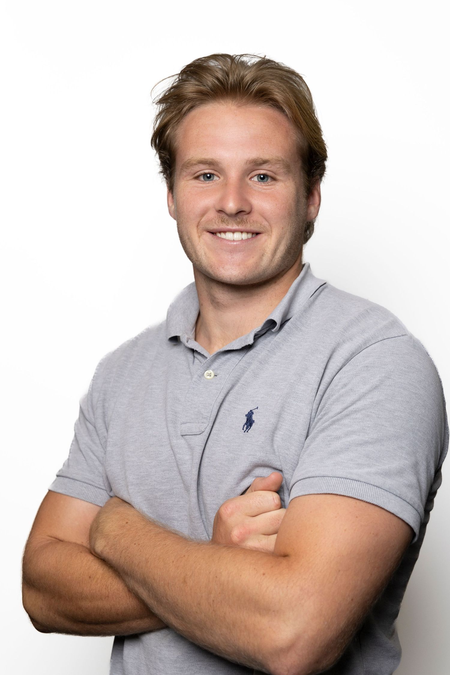 Smiling young man in a gray polo shirt stands with arms crossed against a white background.