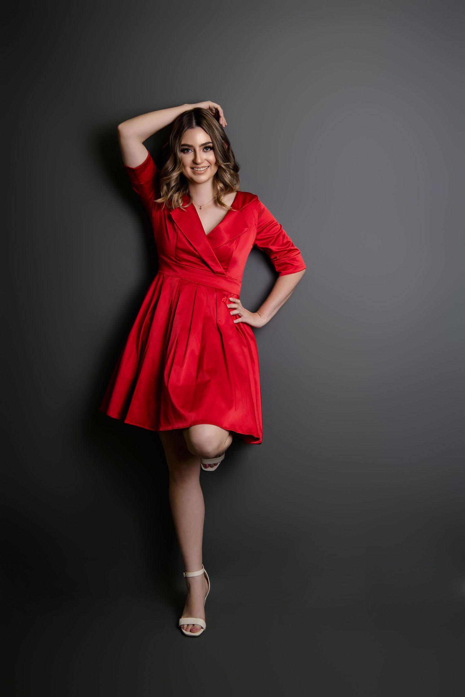Smiling woman in red wrap dress posing against dark gray wall in strappy heels.