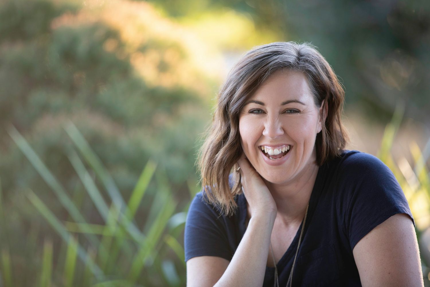 Happy woman with short wavy hair laughing outdoors among green plants in natural light.