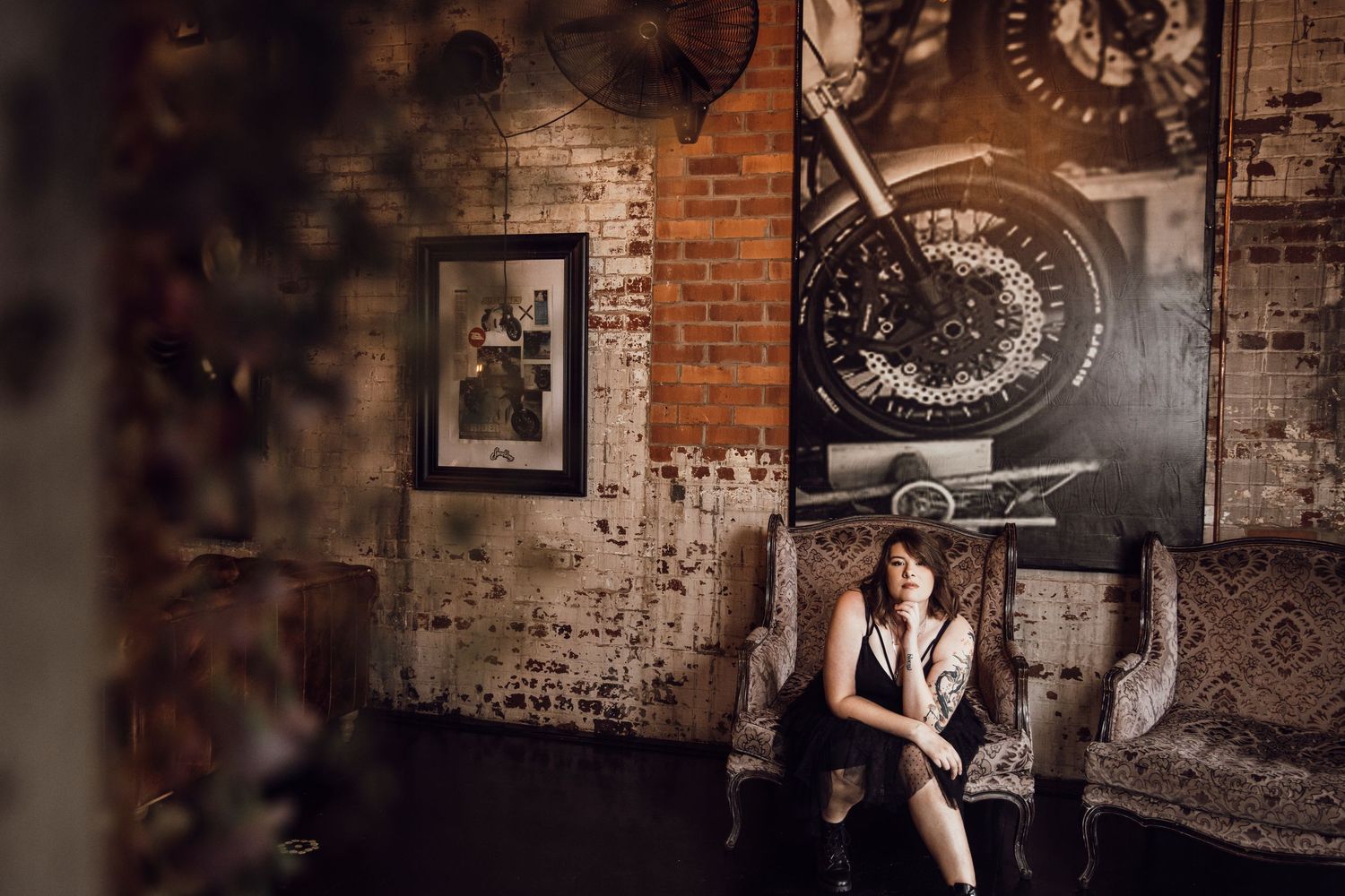 Woman in black outfit sits in moody industrial space with brick walls and motorcycle art.
