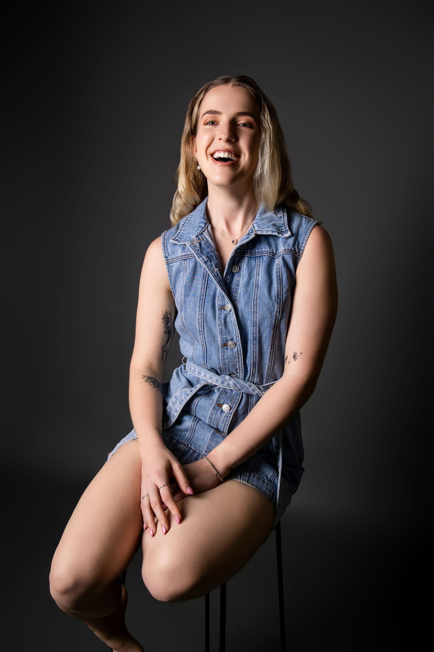 Smiling blonde woman in denim romper sitting on stool against dark gray background.