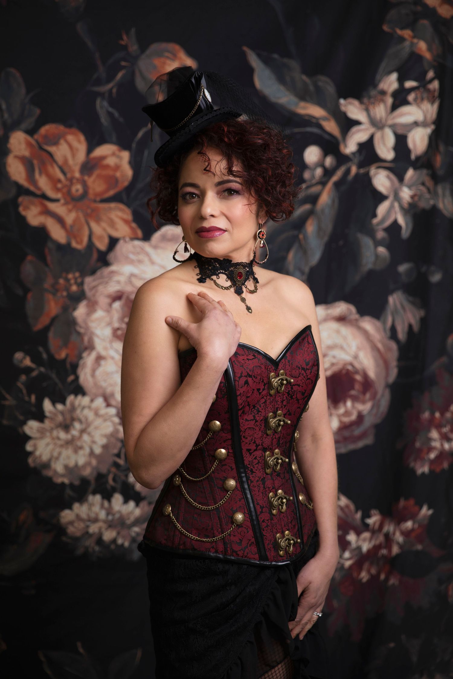 Woman in steampunk costume with red corset, gold accents, black mini top hat, and lace choker against floral backdrop.