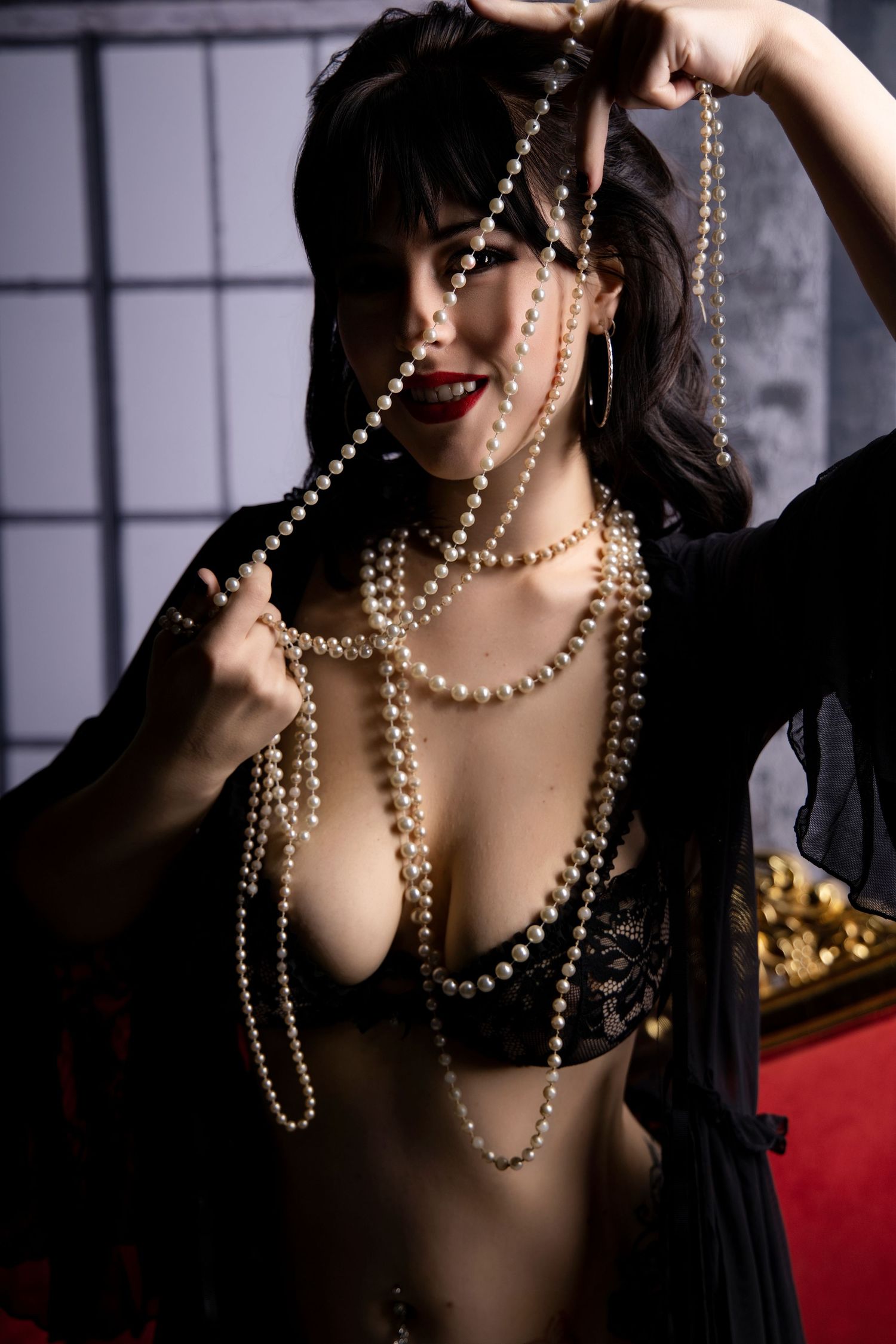 Sultry brunette woman draped in pearl necklaces wearing black lace bra in dramatic studio portrait.