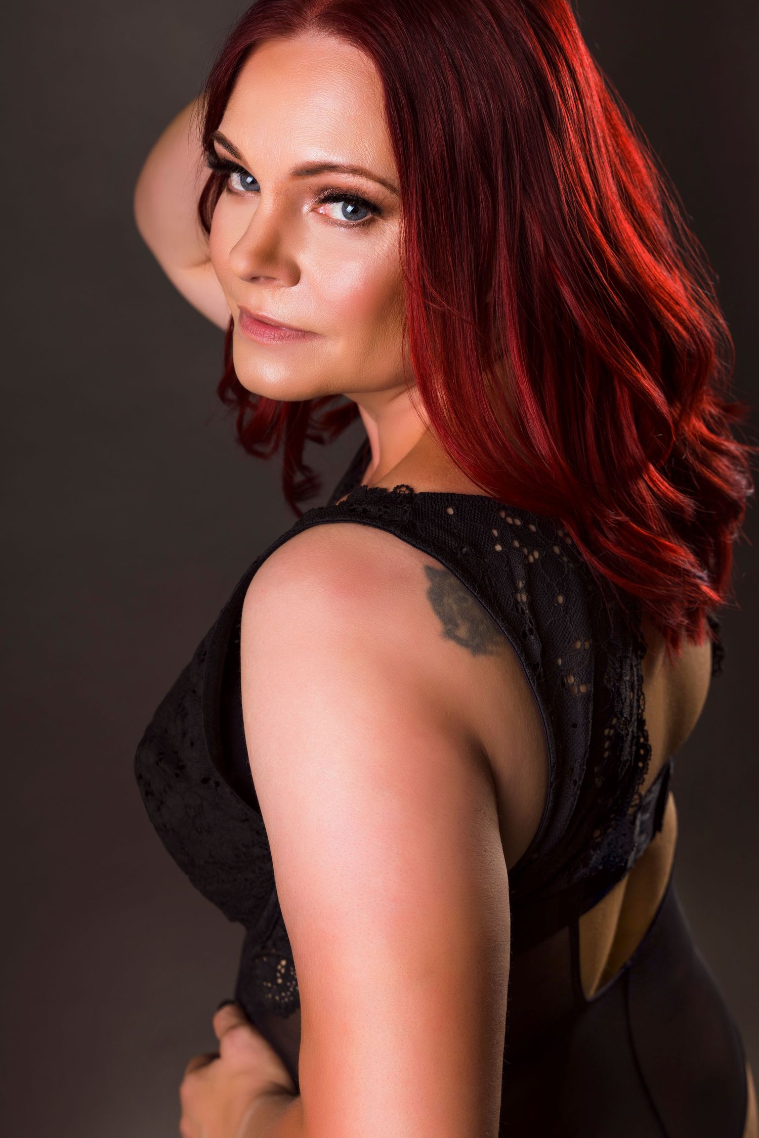 A confident woman with vibrant red hair and a shoulder tattoo poses in a black lace top against a dark background.