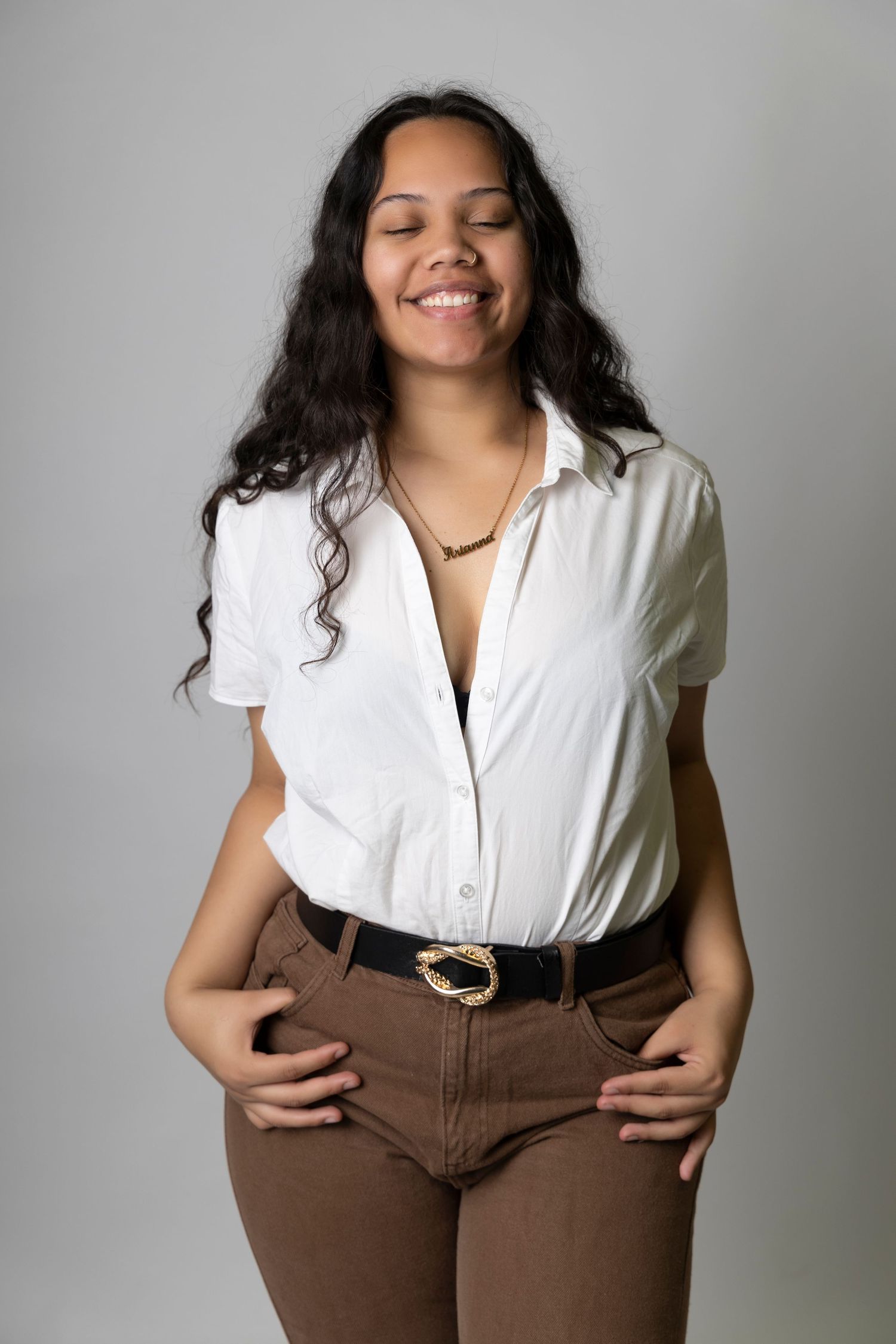 Smiling young woman with closed eyes in white shirt and brown pants on gray background.