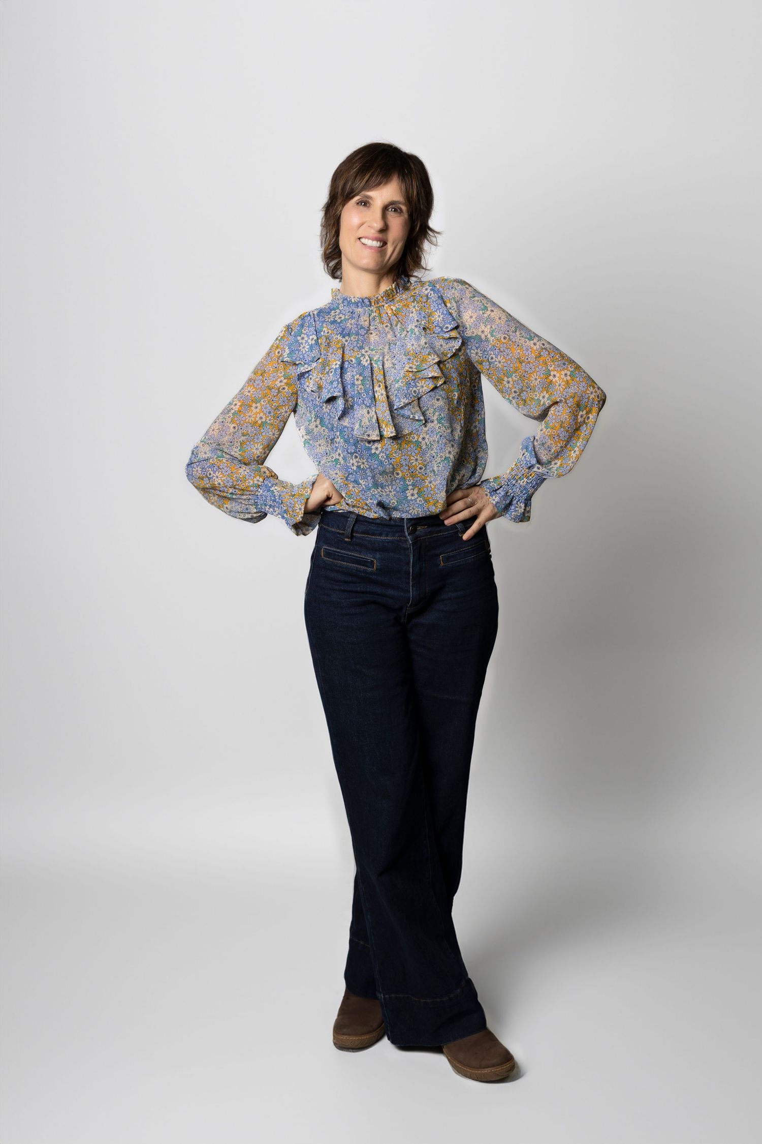Woman in floral ruffled blouse and dark flared jeans standing confidently with hands on hips in white studio.