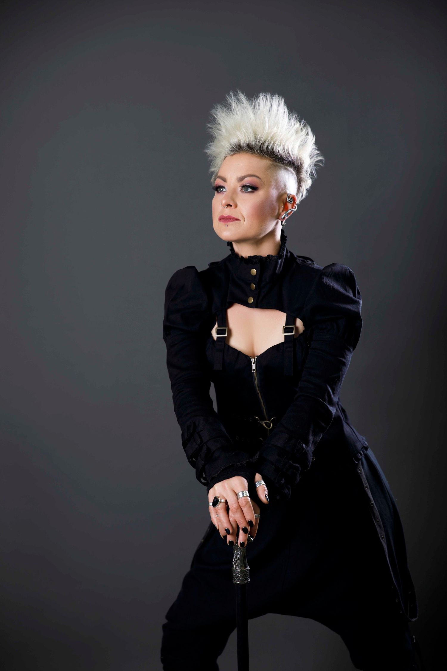 Woman with blonde mohawk wearing black structured coat holding cane in dramatic studio portrait.