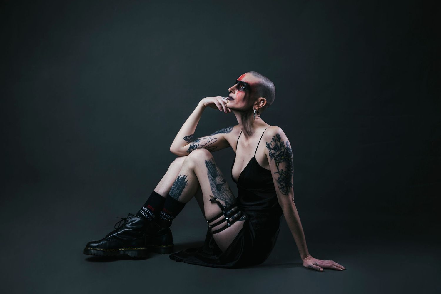 Tattooed woman with short hair sitting on floor in black dress and combat boots on dark background.