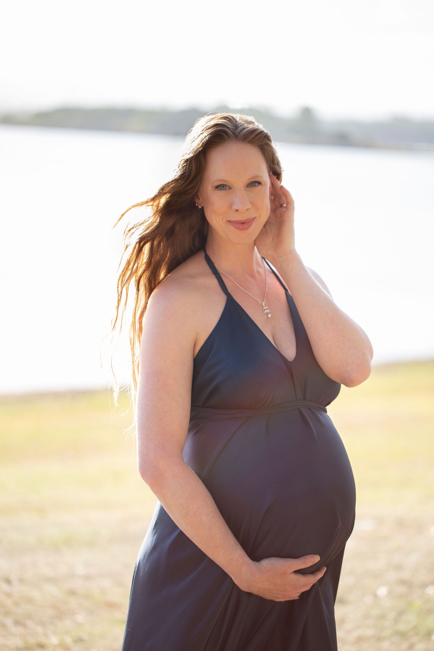 Pregnant woman in navy halter dress holding bump near waterfront during golden hour photoshoot.