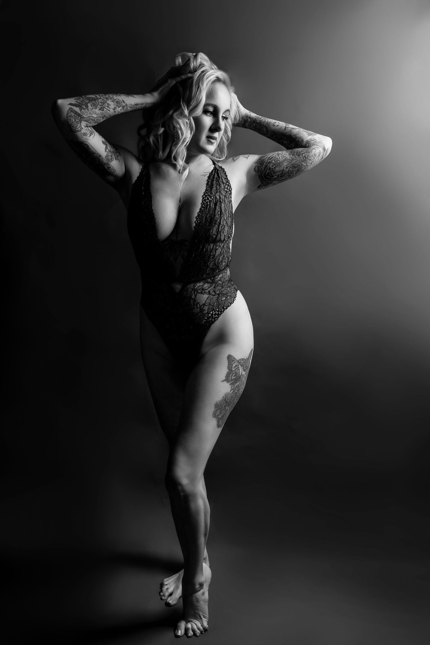 Black and white boudoir photo of tattooed blonde woman posing in lace bodysuit with hands in hair.