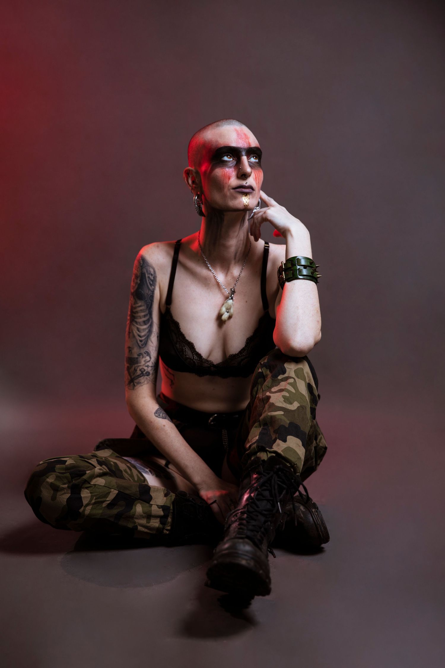 A tattooed bald woman with red face paint sits in camouflage pants and a black bra in a studio.