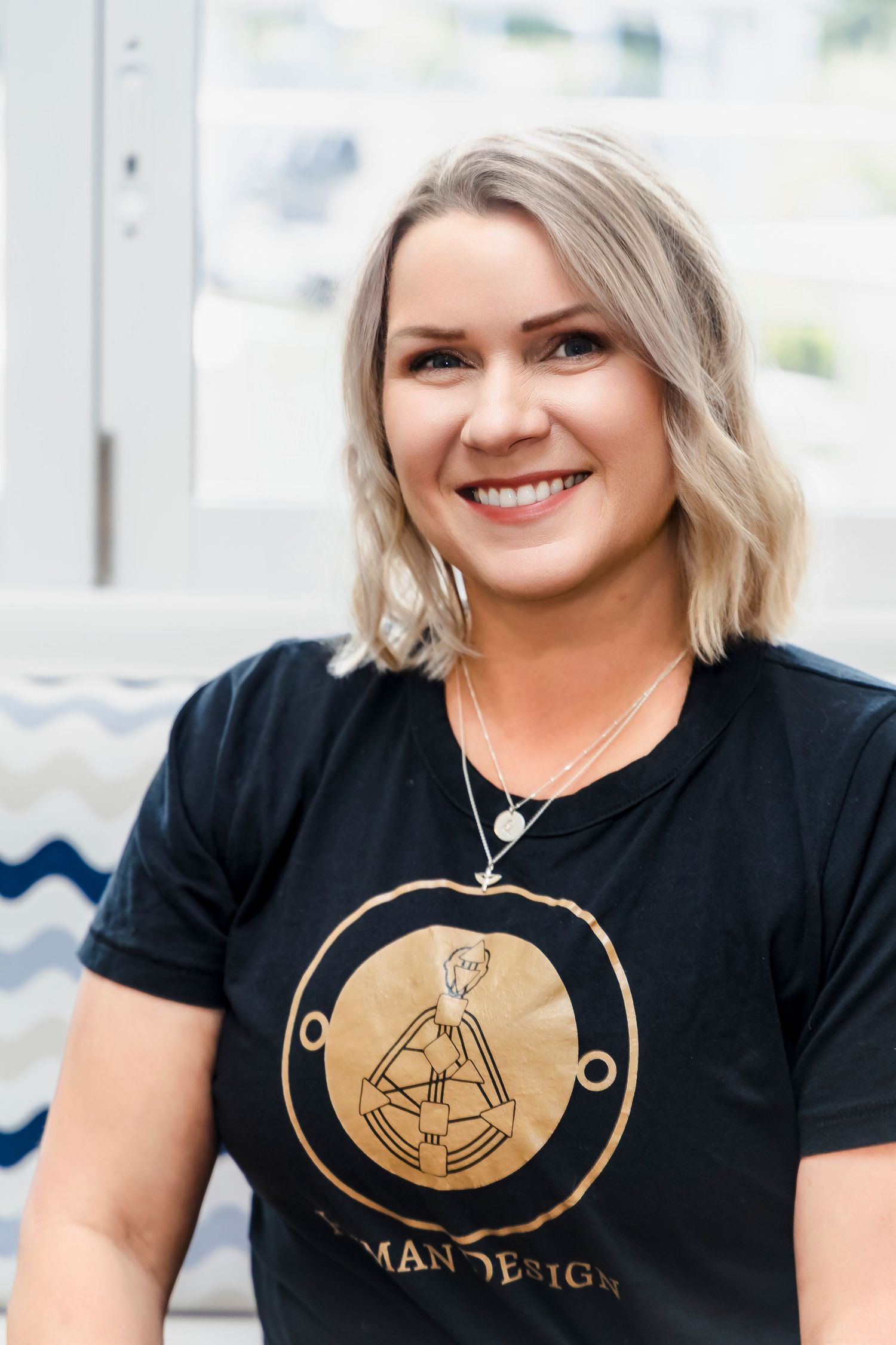 Smiling blonde woman in a black Human Design t-shirt posing near a bright window.