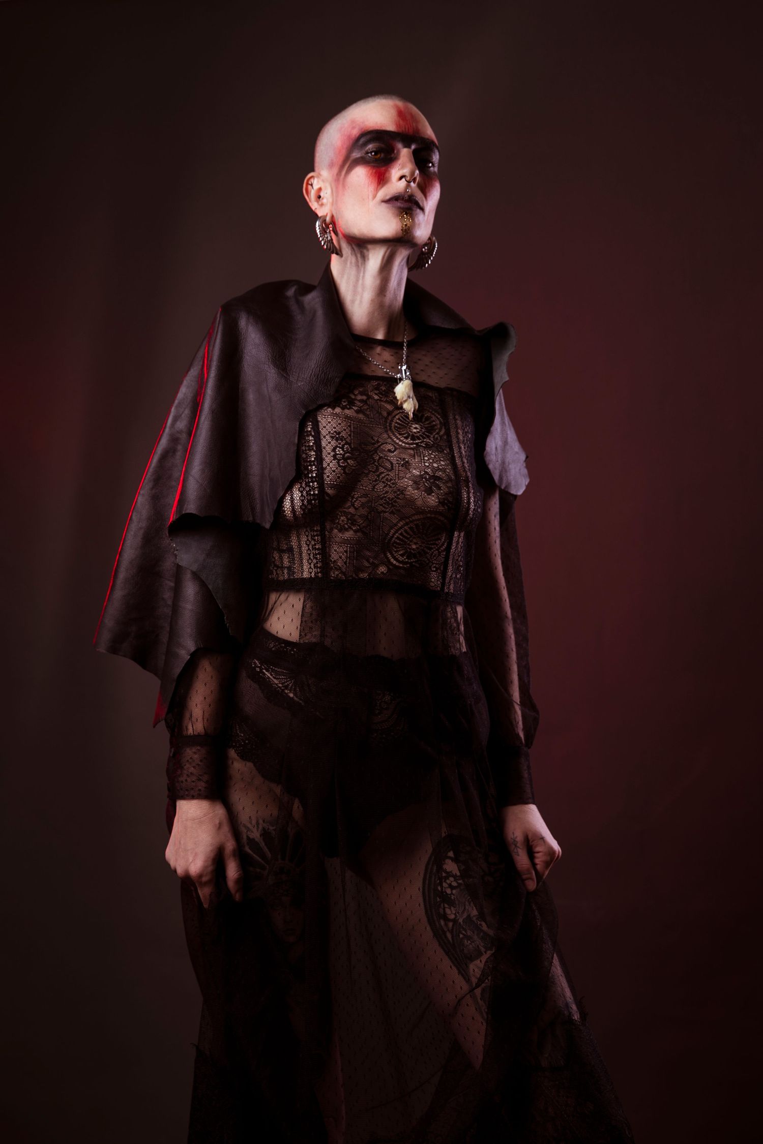 A bald woman with bold red face paint wears a dark lace outfit and cape in a moody studio shot.