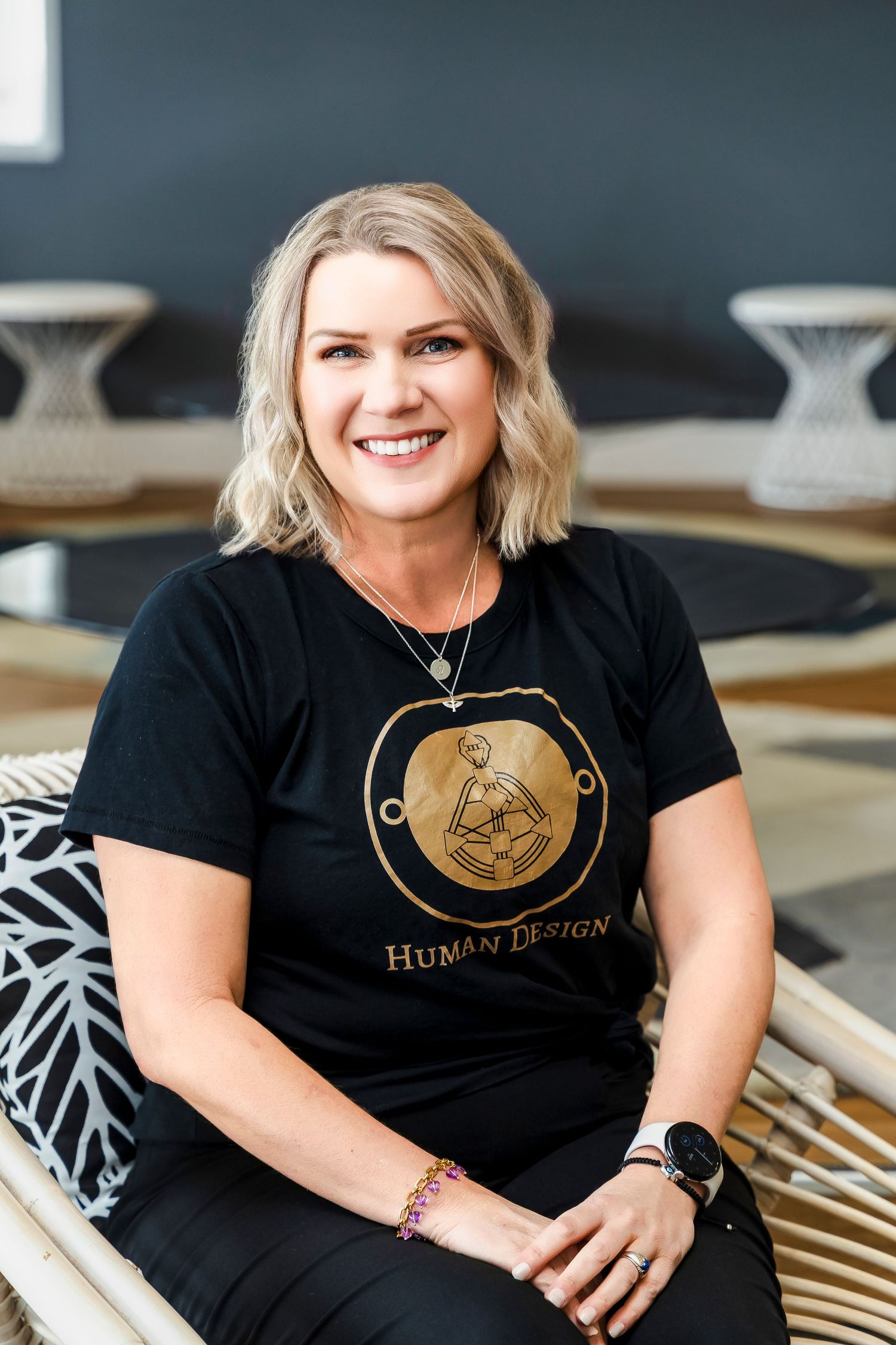 Smiling blonde woman wearing a black Human Design t-shirt seated in a modern interior.