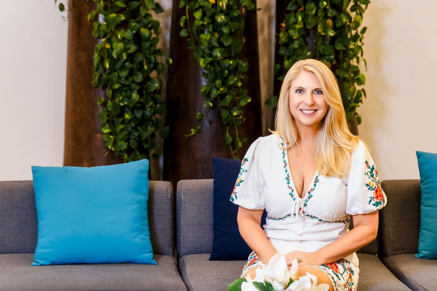 Smiling blonde woman in floral dress seated on a grey couch with blue cushions.