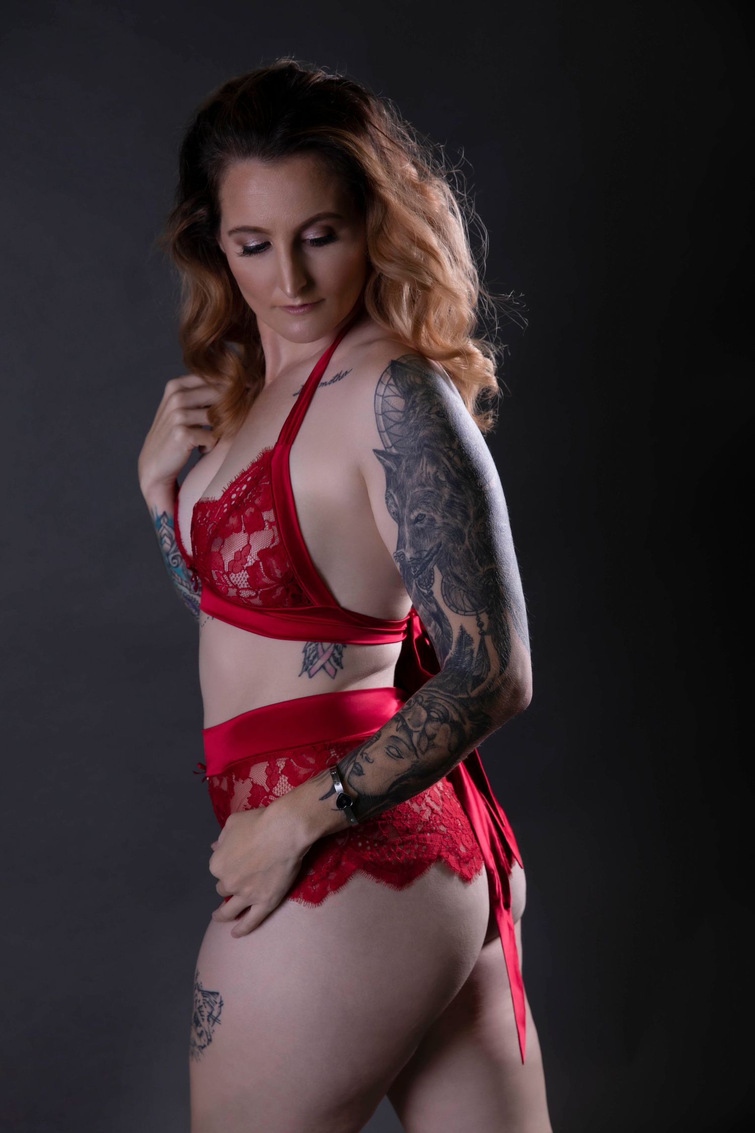 Tattooed woman in red lace lingerie set posing against dark gray studio background.