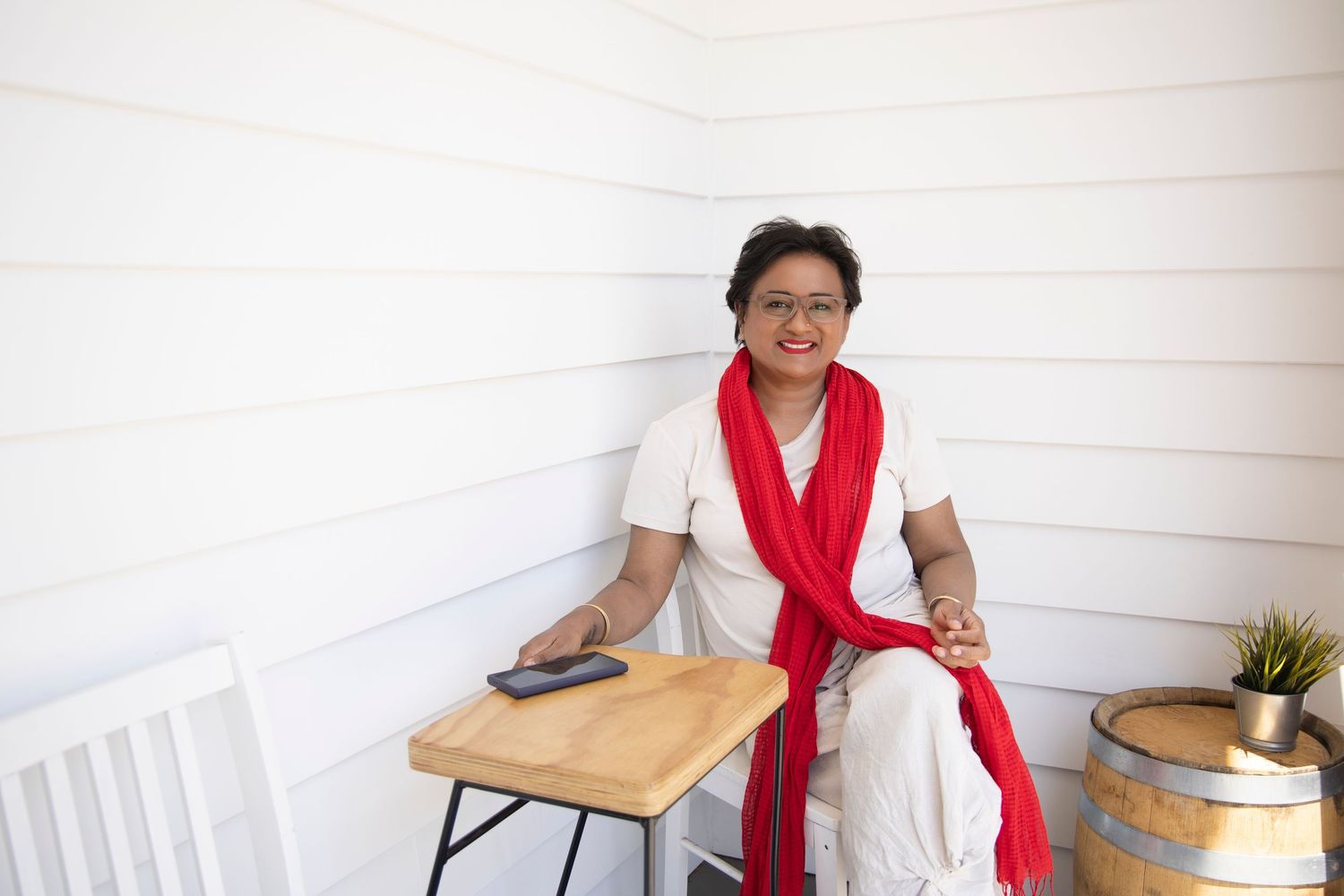 Smiling woman in white outfit with red scarf sitting on porch bench beside rustic wooden barrel.