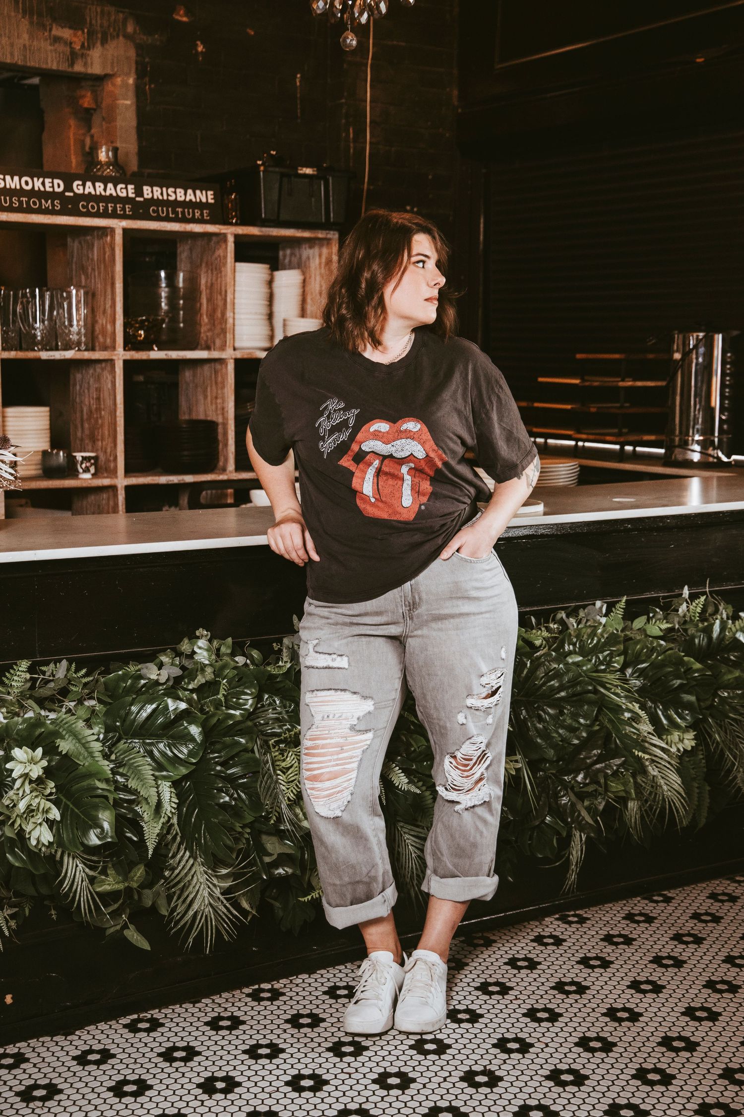 Woman in Rolling Stones tee and ripped jeans poses at a cafe bar counter.