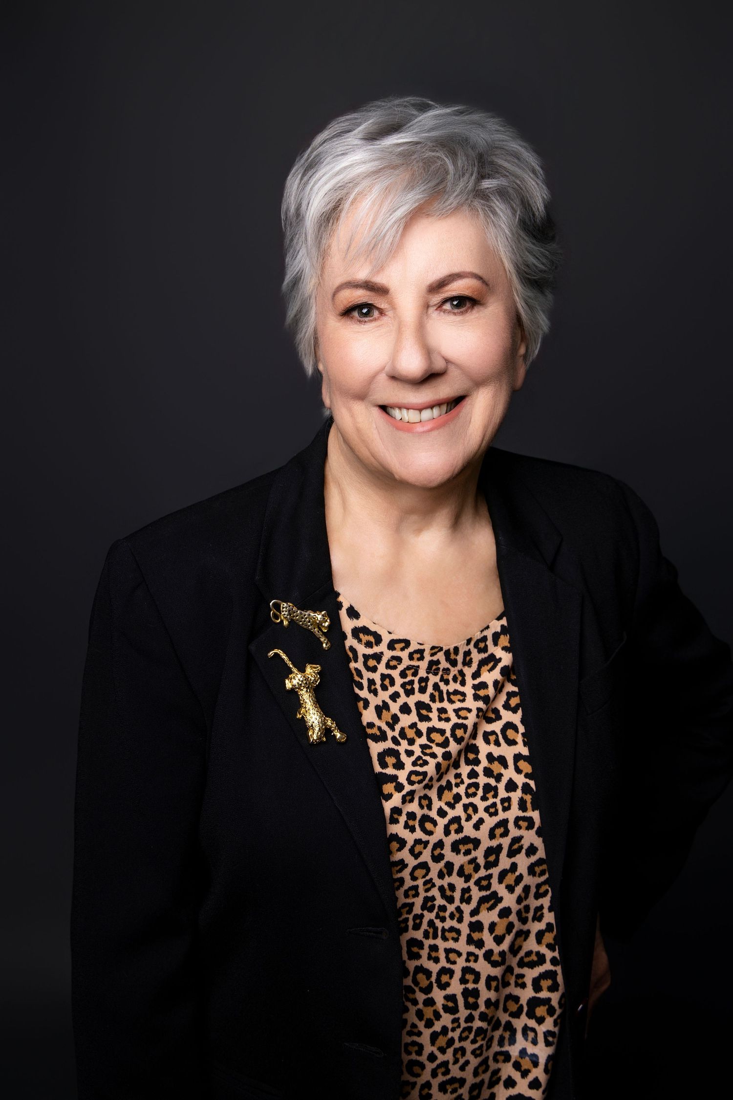 Elegant older woman with short gray hair in black blazer and leopard print top smiling.
