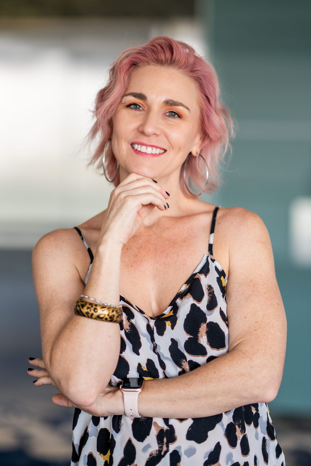 A cheerful woman with short pink hair poses thoughtfully in an animal print dress with a leopard print bracelet.