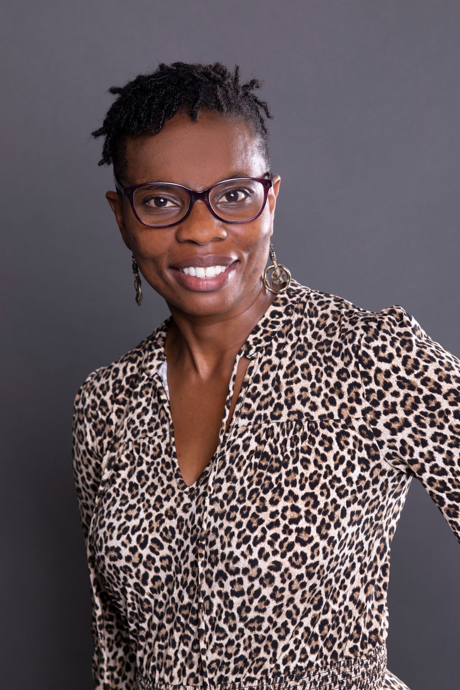 Confident Black woman in leopard print dress and glasses smiling against gray background.