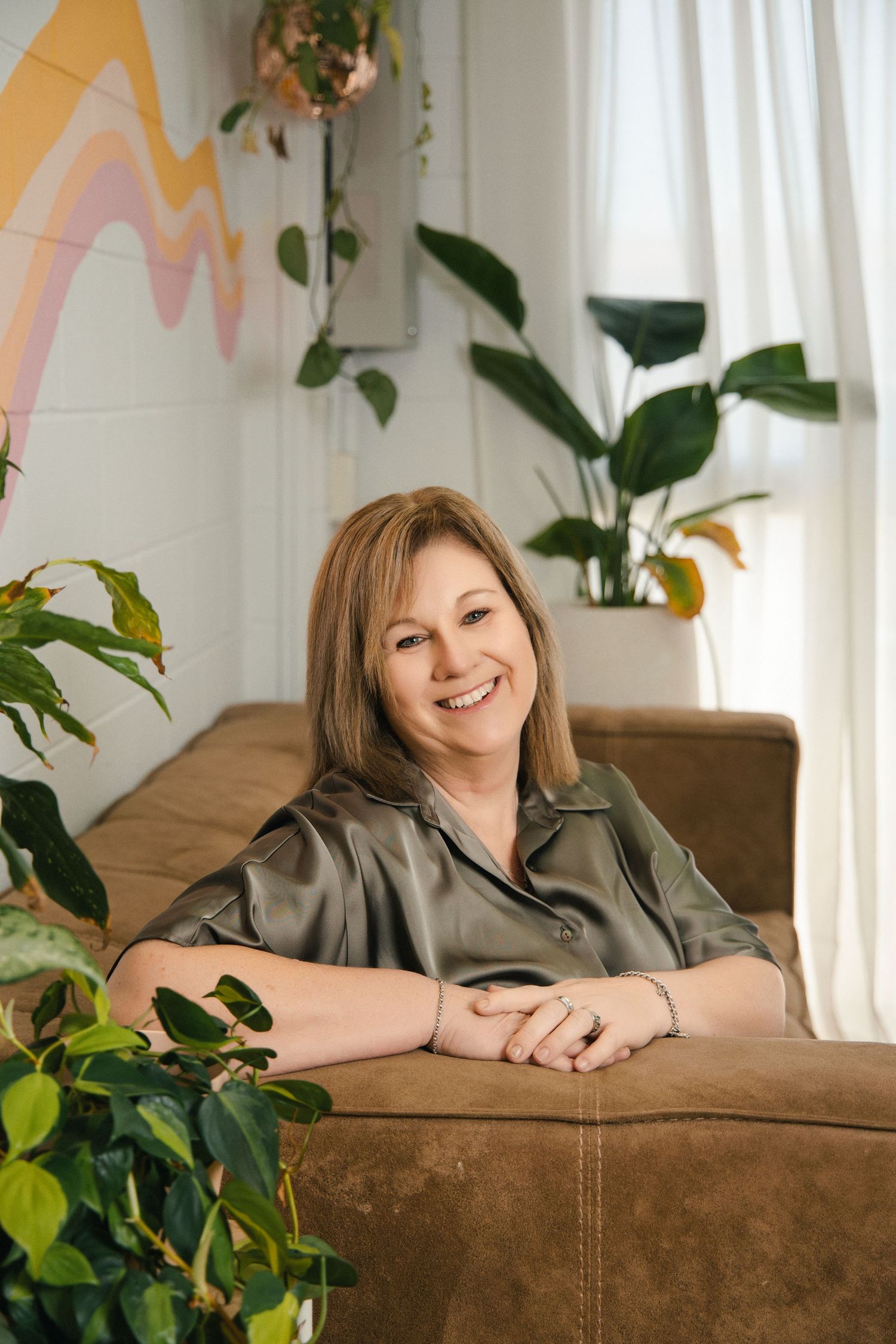 Smiling woman in olive satin blouse seated on brown sofa surrounded by lush indoor plants and colorful wall mural.