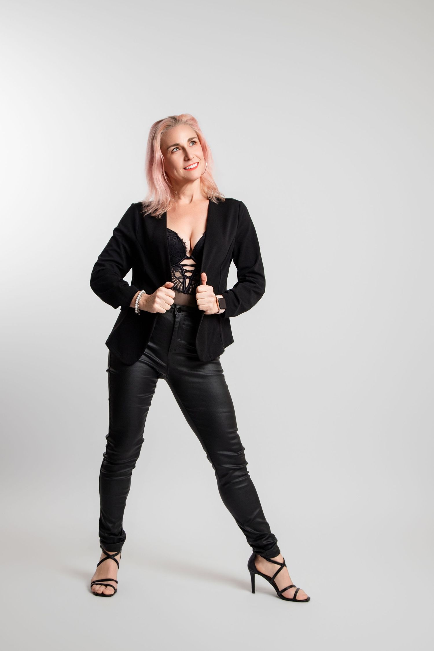 Woman with pink hair in black blazer and leather pants posing in white studio.