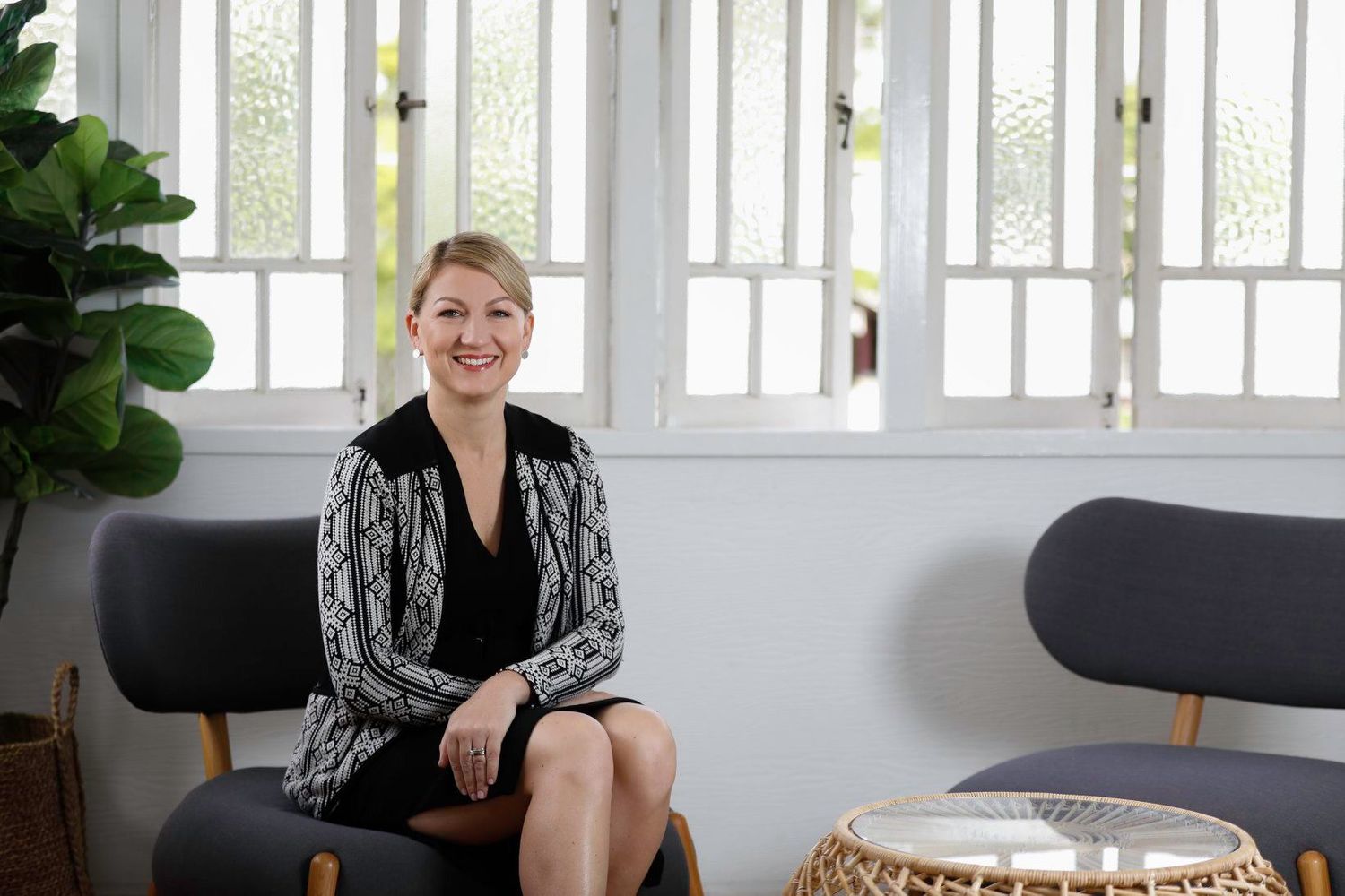 Professional woman in black dress and patterned blazer sitting in modern office with white shuttered windows.