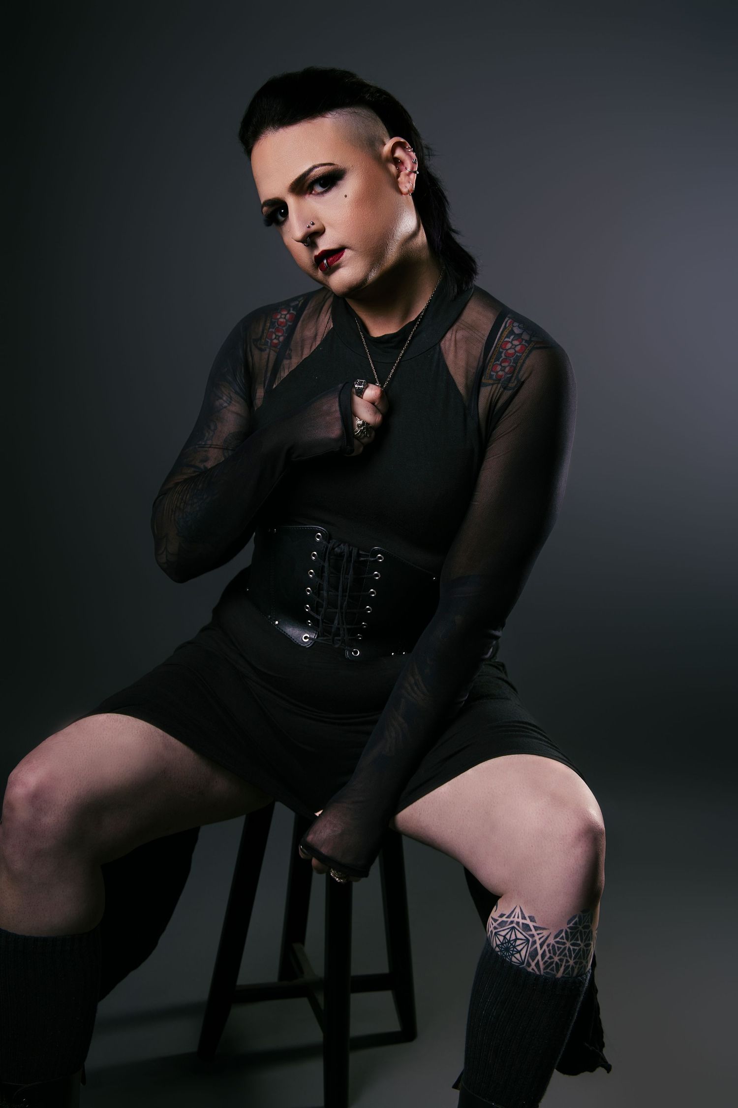 Gothic woman in black lace top and corset sitting on stool in studio.