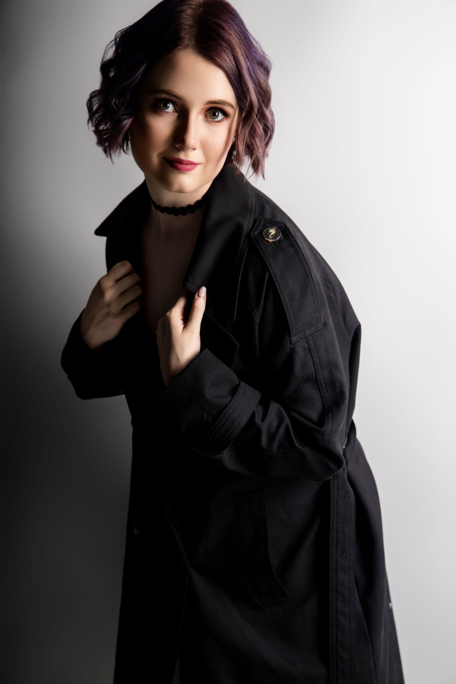 Woman with purple hair in black trench coat posing against gradient gray studio background.