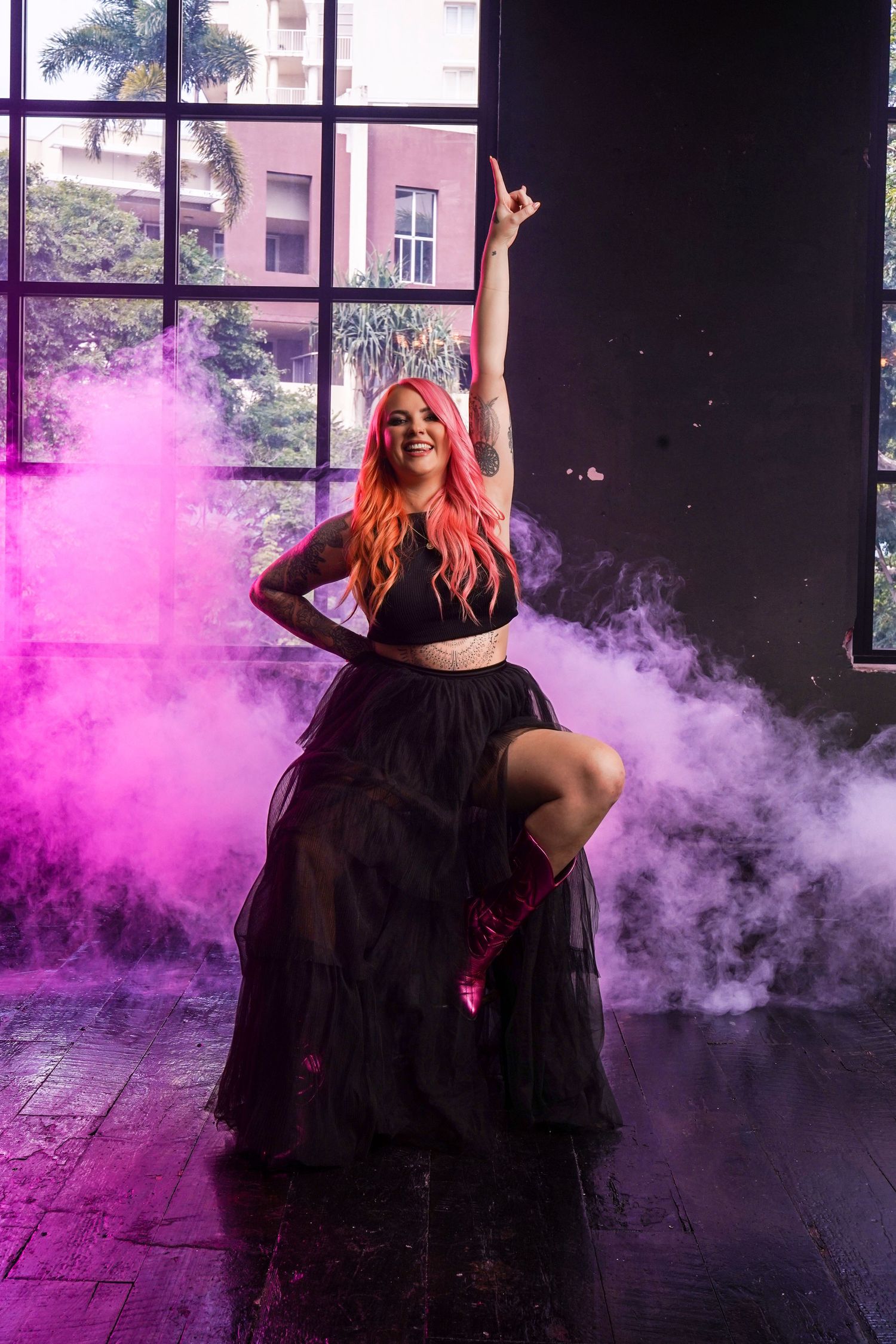 Pink-haired woman in black tulle skirt poses dramatically with pink smoke in studio.
