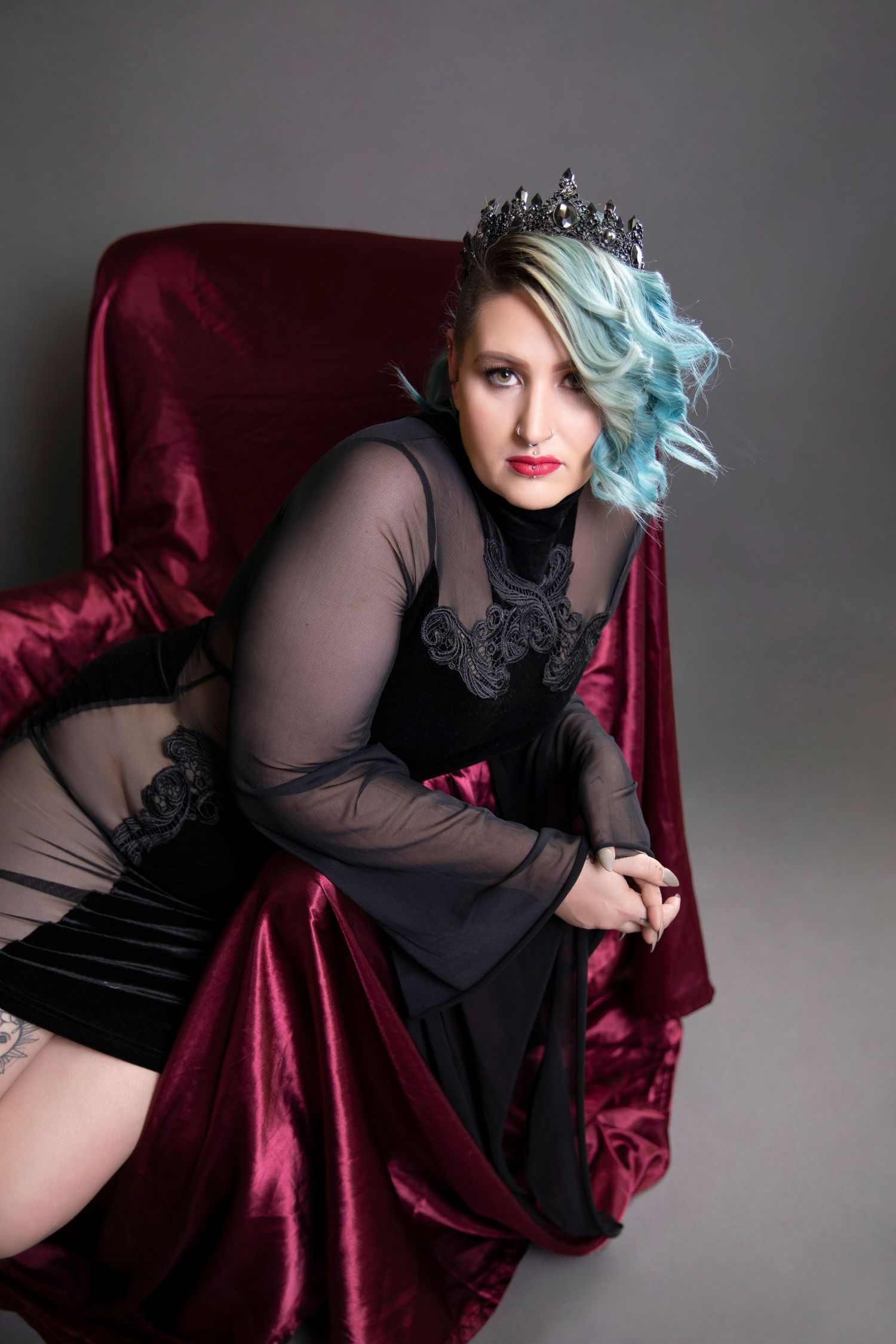 Woman with blue hair wearing black sheer dress and dark crown sits on red velvet chair.
