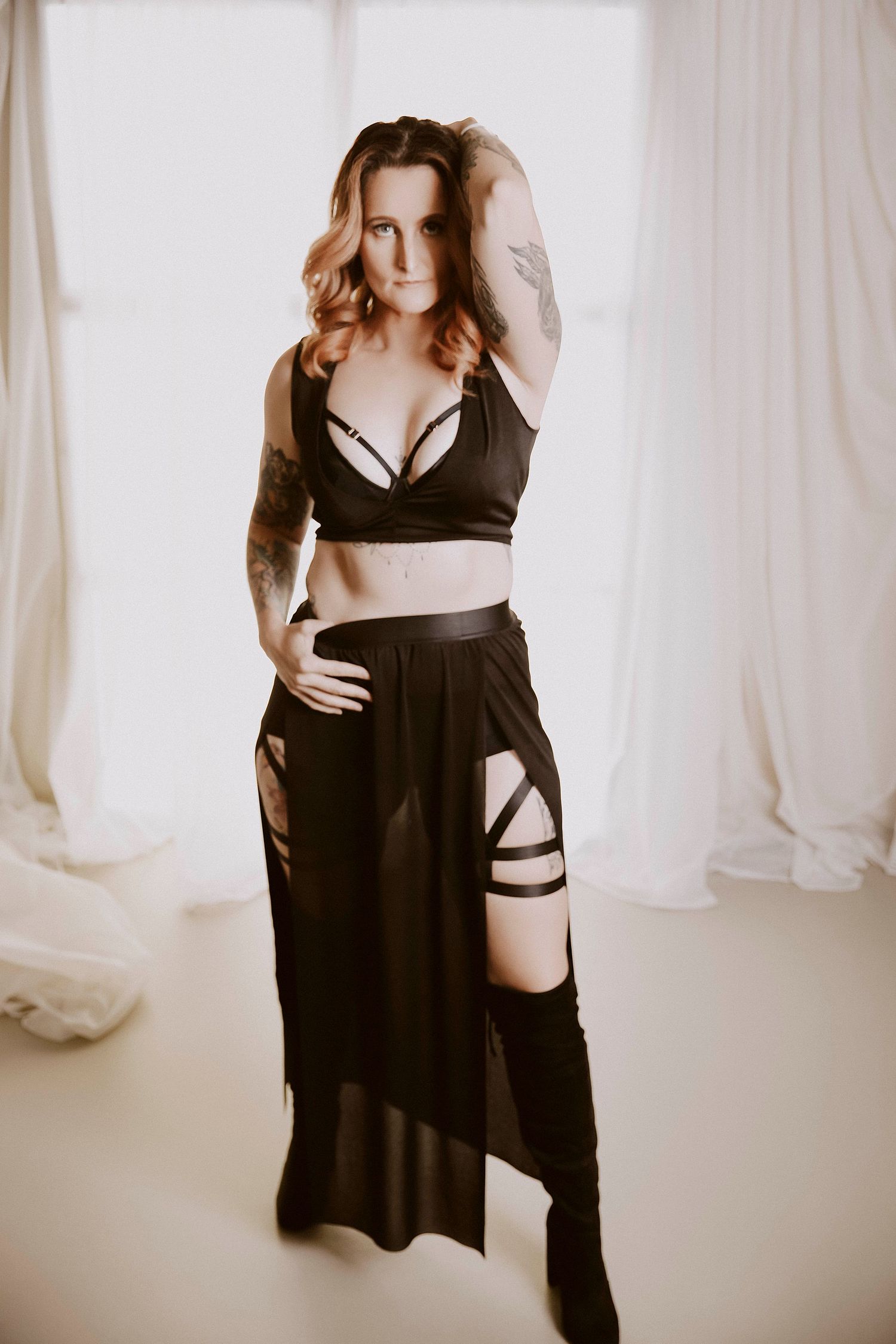 Woman in black crop top and strappy skirt with thigh-high boots posing in bright white room.