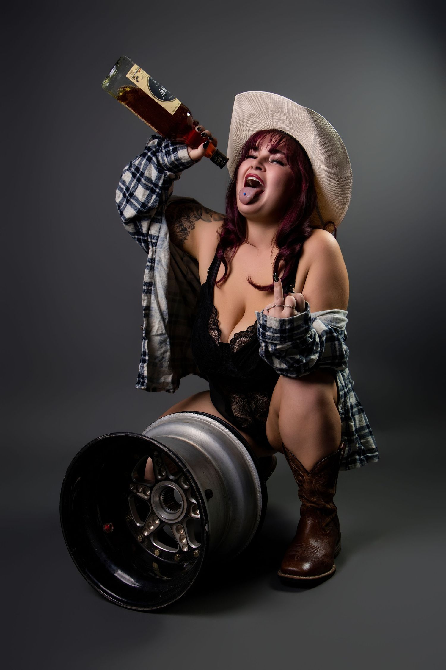 Woman in cowboy hat and lingerie posing with whiskey bottle and car rim in studio.